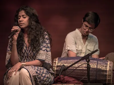 On the left, a woman with long wavy dark hair and patterned blue blouse sings into a microphone, seated, eyes closed. On the right, another person sits behind a long barrel-shaped drum, holding a hand drum, looking down.