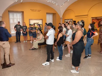 A man in a navy blue shirt and khakis leads a large group of students through an art gallery