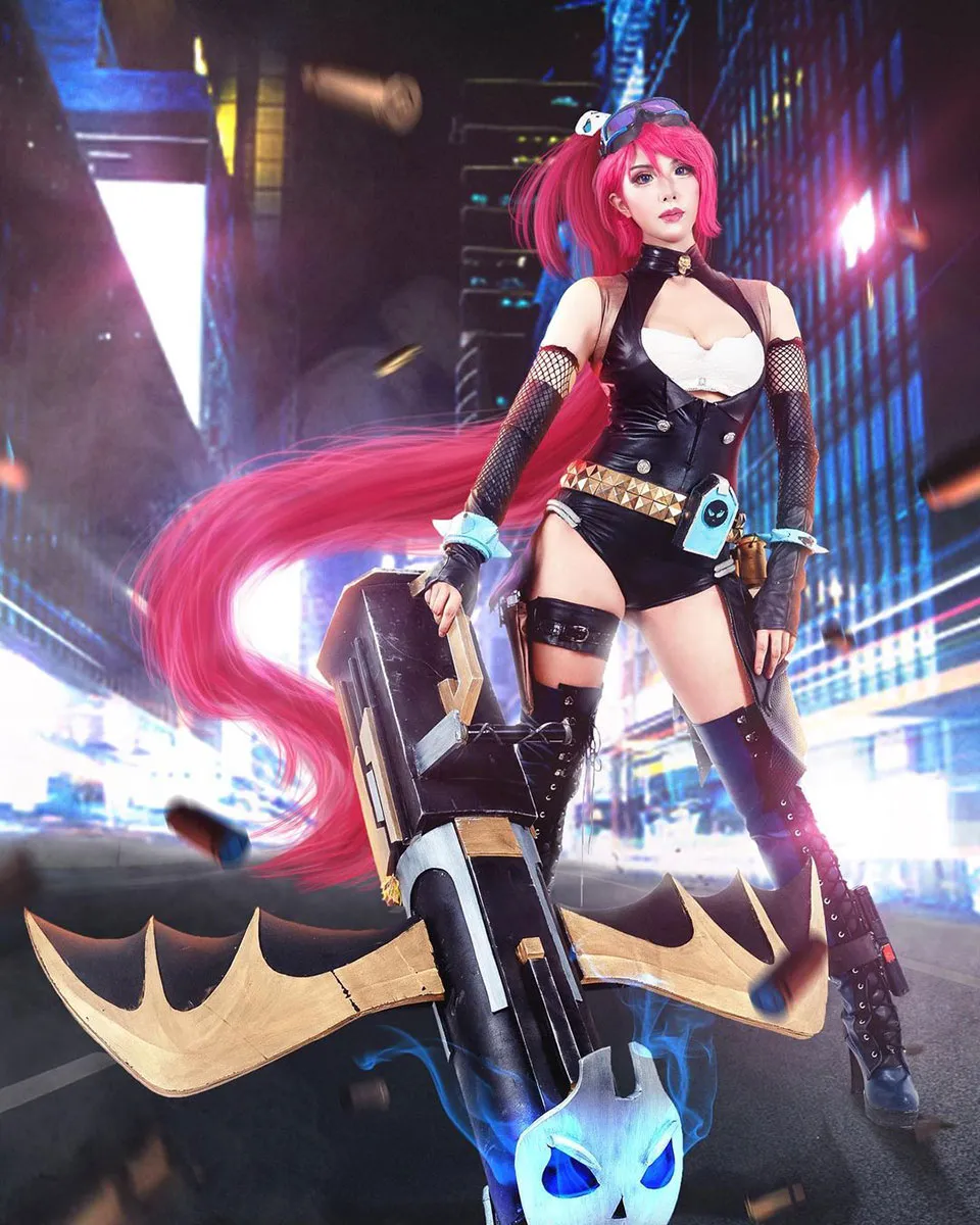 A person poses in a costume of very long windswept pink wig, goggles, black bodysuit, and black combat boots, holding some kind of rocket launcher. The costume is styled to look like a video game rendering, with a bright cityscape in the background.