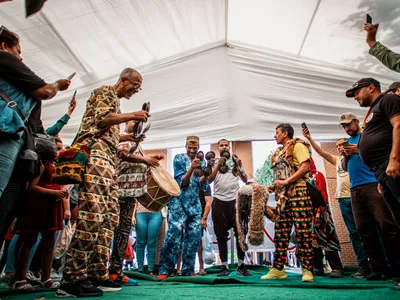 Four men stand together on a tented stage playing percussive instruments. They are dressed in brightly patterned clothes and are filmed by people holding cellphones.