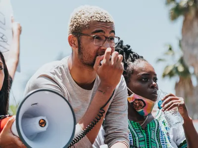 Three young activists are outside at a protest – looking off into the distance. They all appear to be African American. One is holding a bullhorn and has a determined look on her face. Another is holding the bullhorn’s microphone and speaking into it.